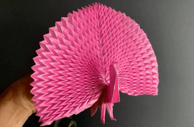 Photo of a pink origami Peacock. The Peacock is being held my a barely visible hand. The background is grey.