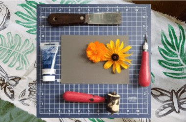 Photo of printmaking Supplies including linoleum, roller, carving tools and paint. There are also two flowers in the photo, one yellow and one orange. There is a background image of green leaf prints.